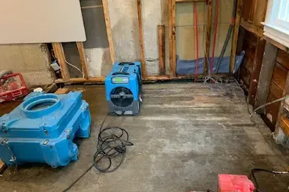 Air movers drying a stripped floor beside open wall framing